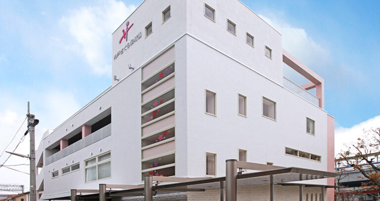 白とピンク色の4階建て介護施設建物の外観。正面にロゴ看板あり。