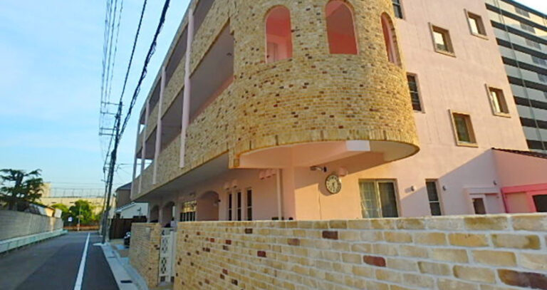 特徴的な建築デザインの介護施設外観。クリーム色のタイル張り壁面と赤いアクセント。
